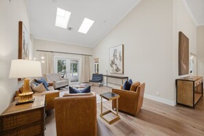 A spacious living room with a sofa, armchairs, a coffee table, and a side table. There is a large window with curtains, a door leading outside, and a painting on the wall.