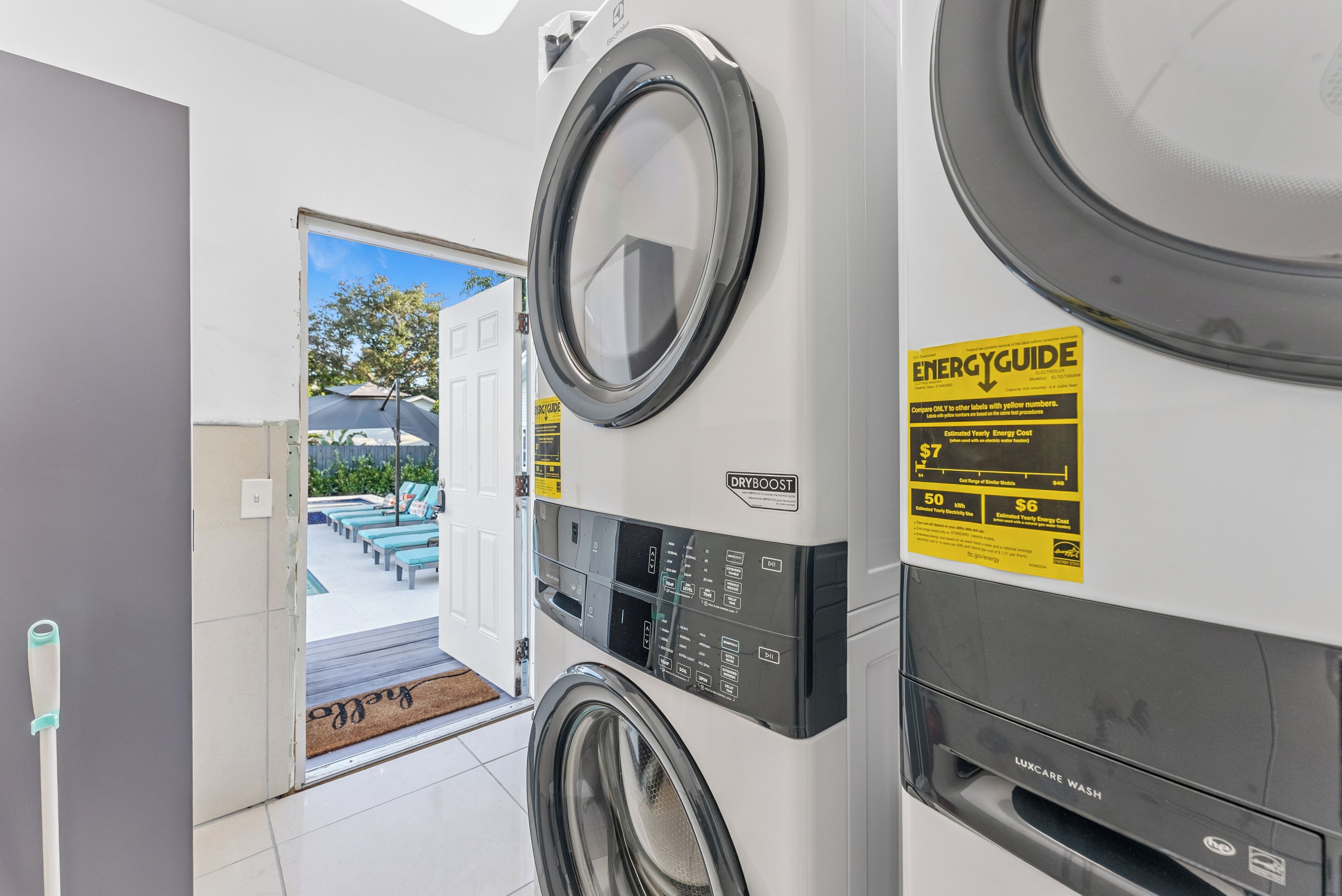 A laundry room with two front-loading washing machines, a control panel, and a door leading to an outdoor area with a pool and lounge chairs.
