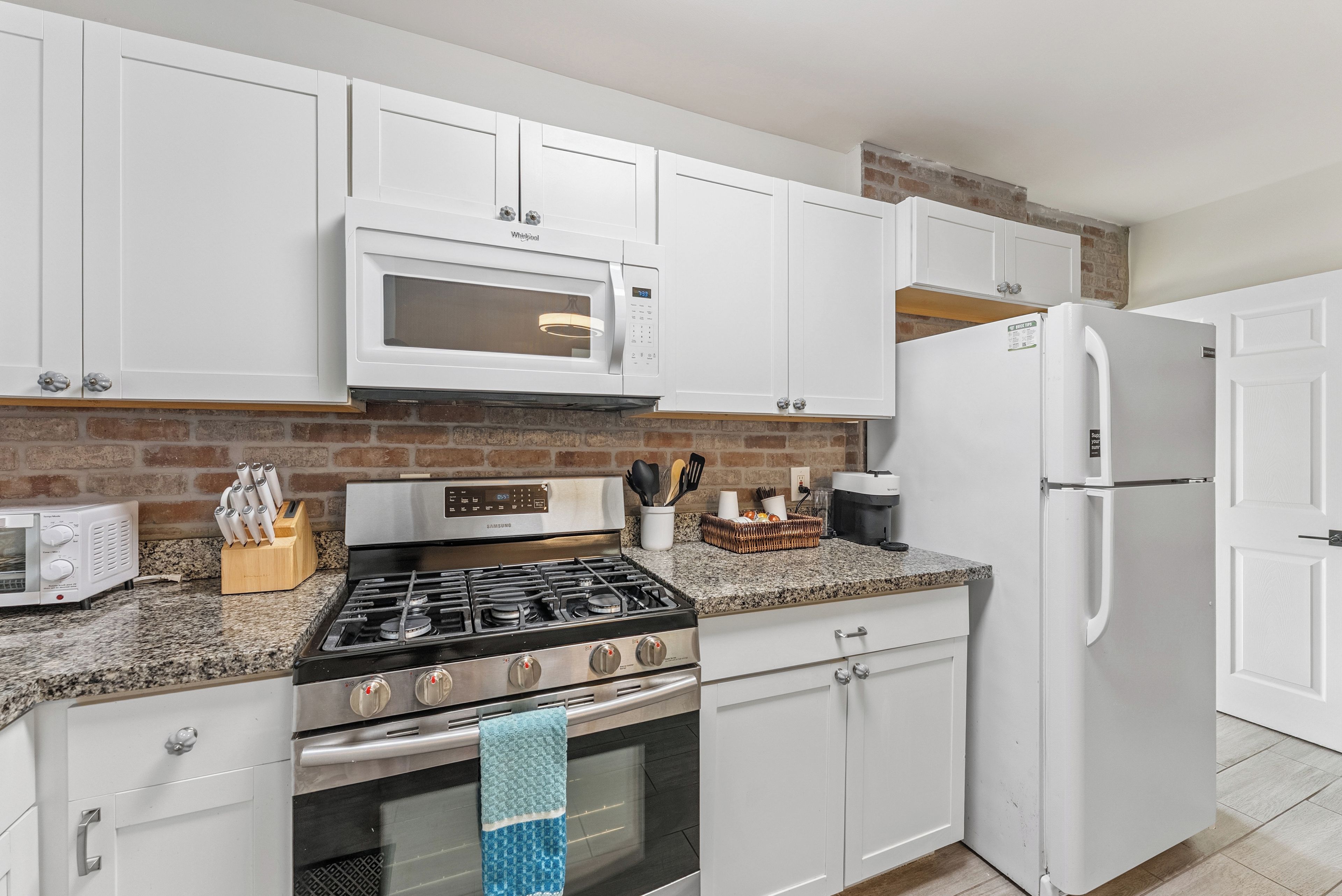 A kitchen with white cabinets, a microwave, a gas stove, a refrigerator, and a brick wall.