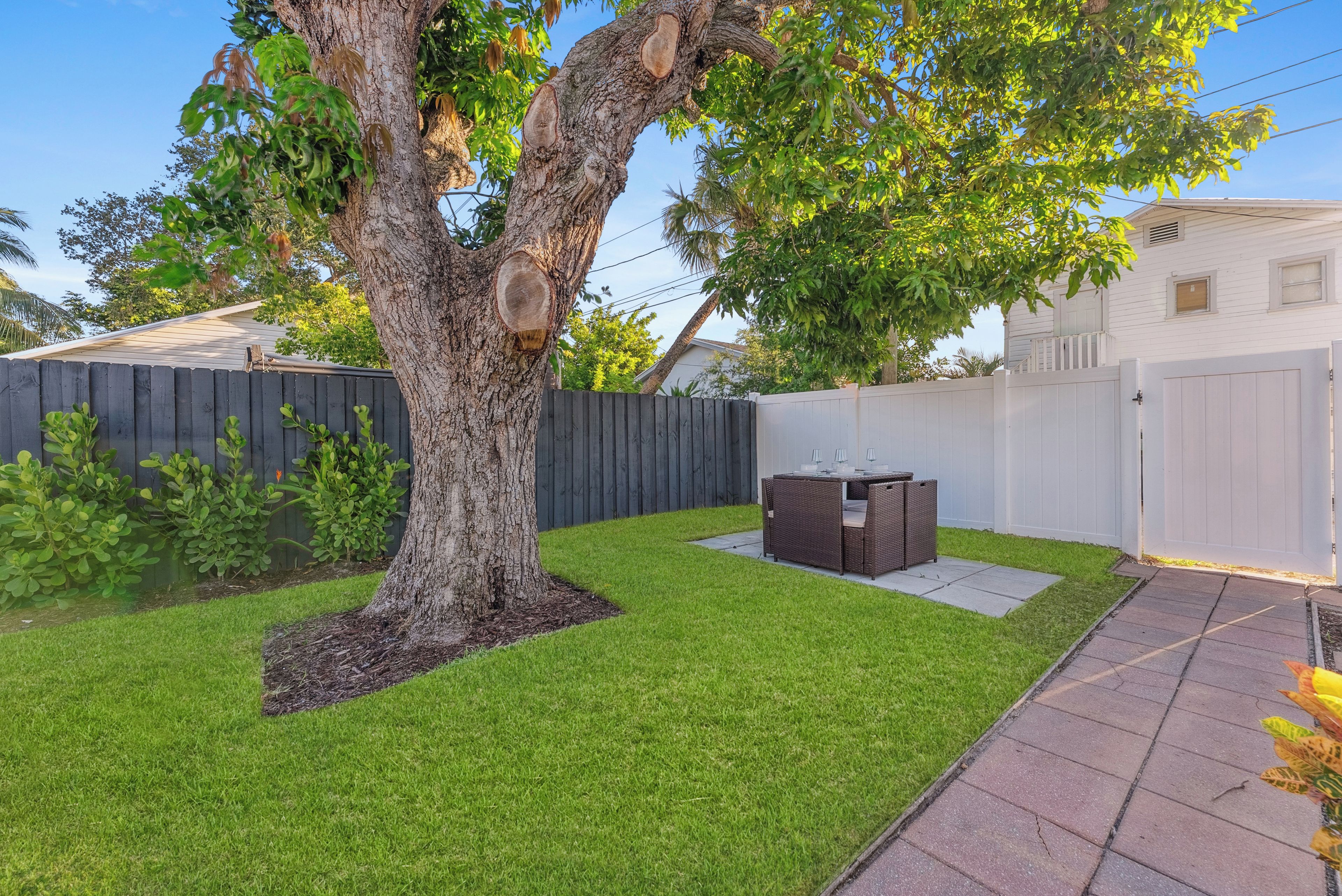 A backyard with a tree, a patio table, and a white fence.