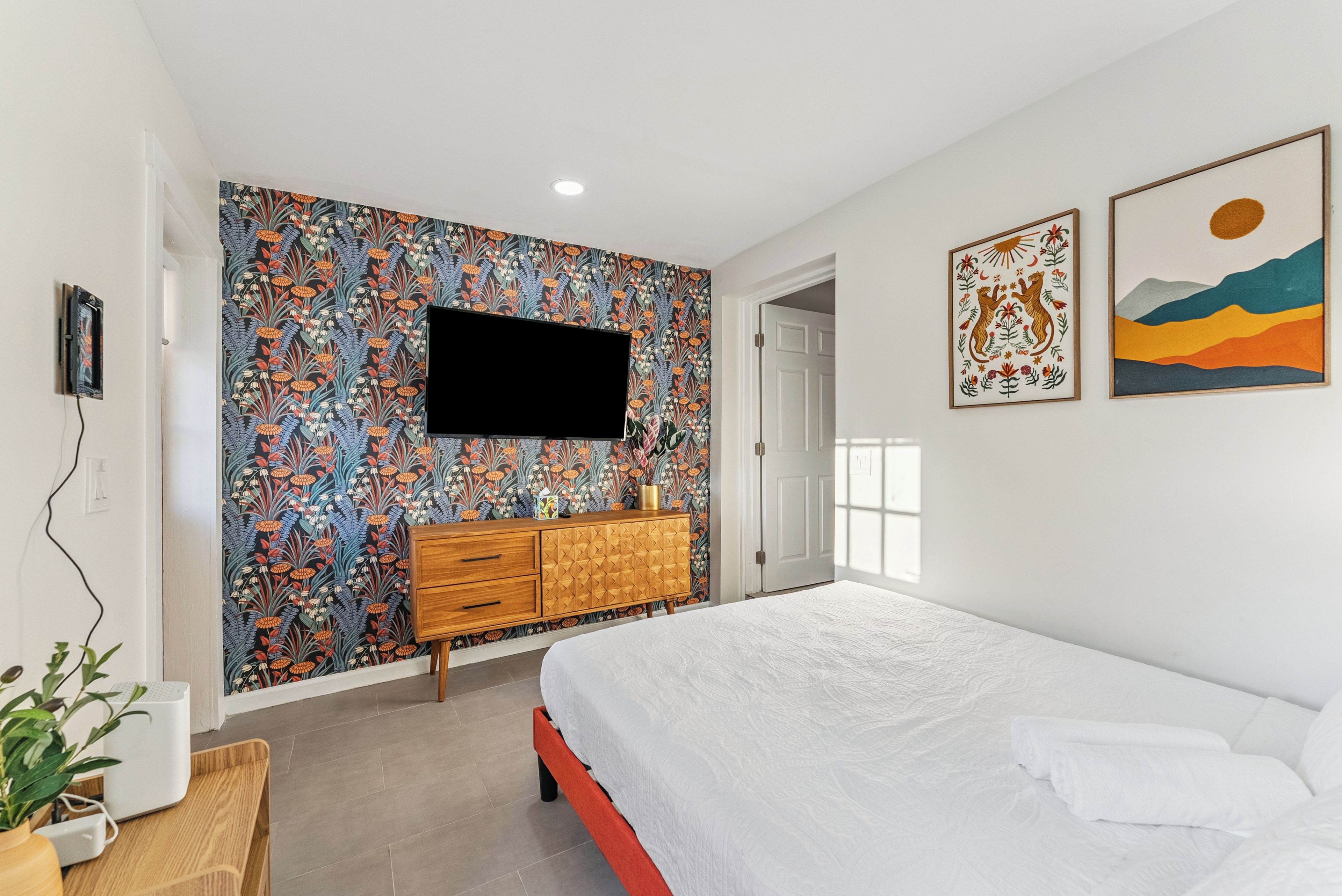A bedroom with a bed, a TV mounted on a floral wallpaper wall, a wooden dresser, and two framed artworks on the wall.