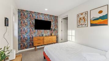 A bedroom with a bed, a TV mounted on a floral wallpaper wall, a wooden dresser, and two framed artworks on the wall.
