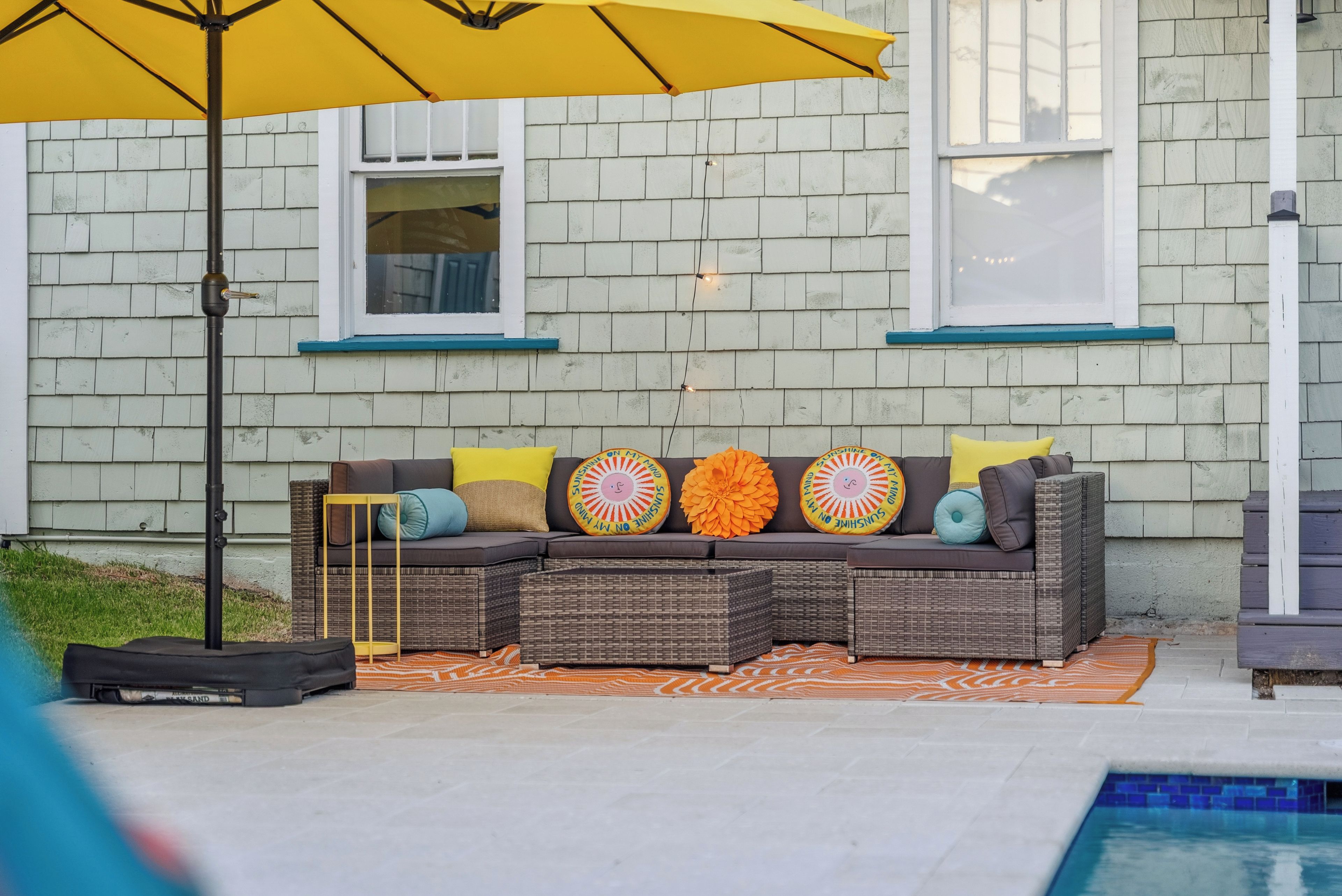 A patio area with a wicker sofa, colorful cushions, a yellow umbrella, and a pool.