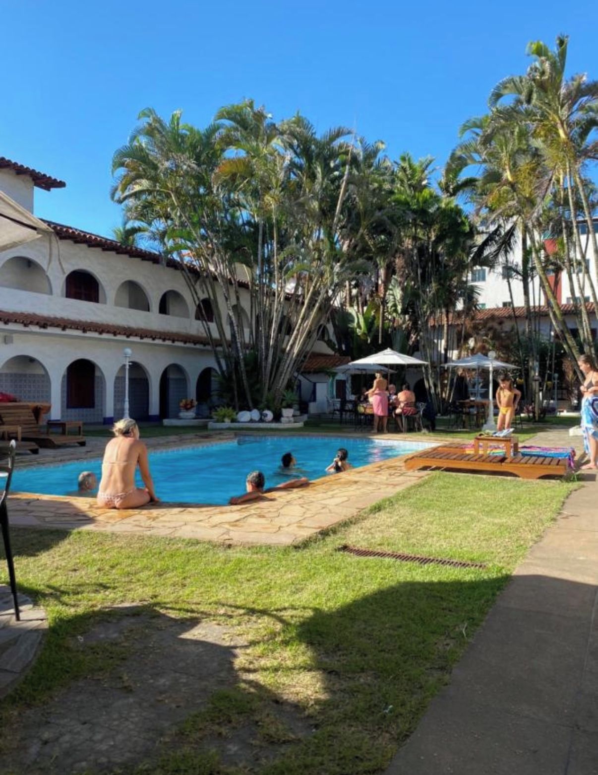 A pool area with people relaxing, surrounded by palm trees and a building with arches.