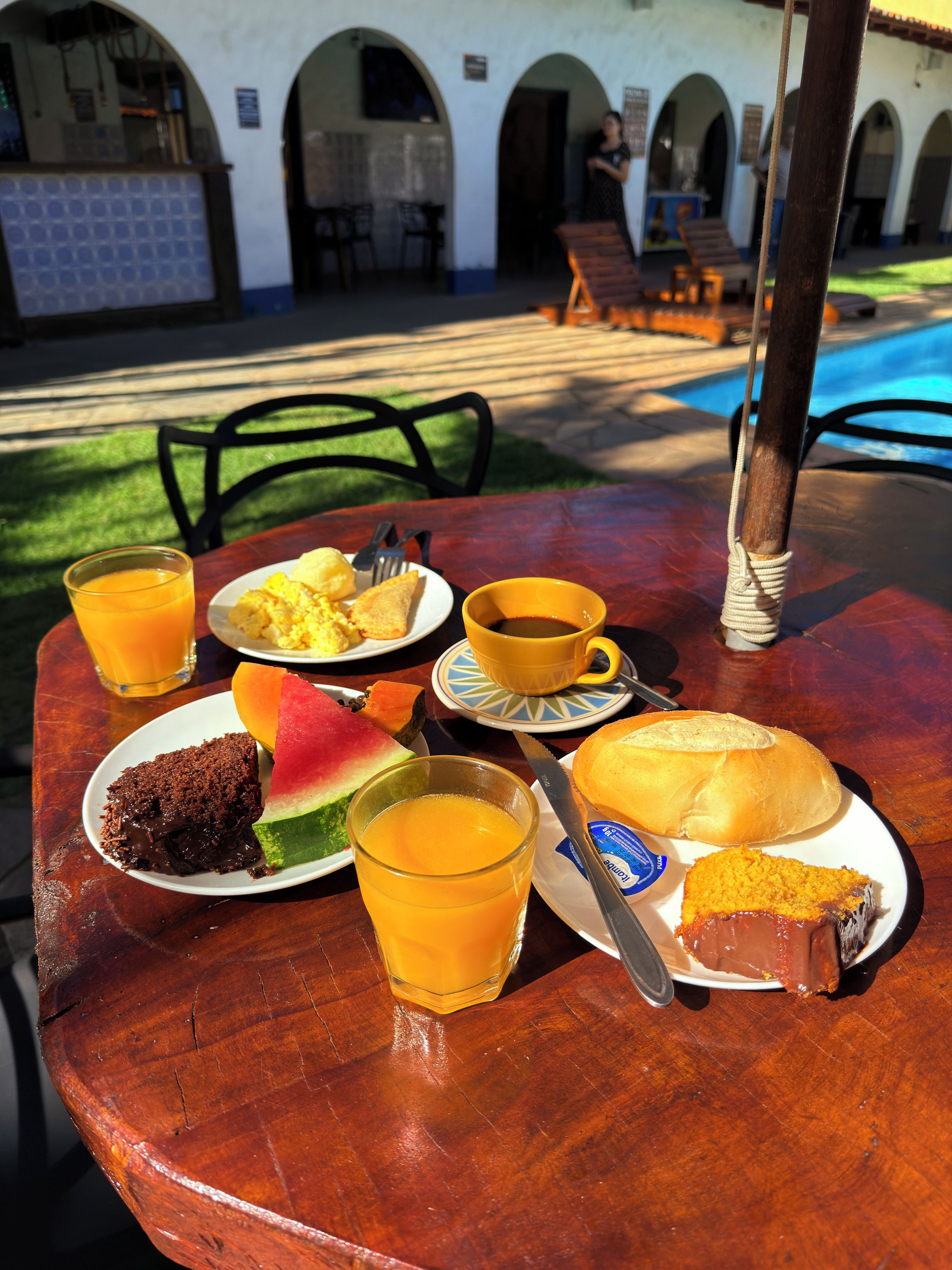 Una mesa con alimentos para el desayuno, incluyendo una porción de panqueques, una rebanada de pastel, un vaso de jugo de naranja, una taza de café y una tajada de sandía.