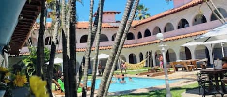 Un área de piscina con palmeras, un edificio con arcos y mobiliario exterior.