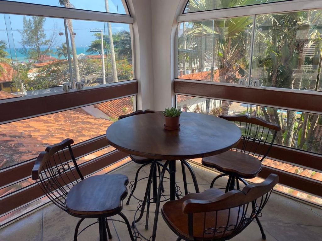 Mesa redonda com quatro cadeiras e um vaso de planta, disposta numa varanda com vista para uma praia e palmeiras.