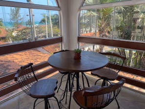 A round table with four chairs and a potted plant, set on a balcony with a view of a beach and palm trees.