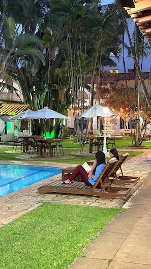 A poolside area with lounge chairs, umbrellas, and a person reading a book.