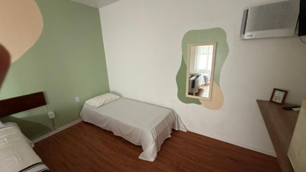A single bed with a white sheet in a room with green walls, a small mirror, and a wooden floor.