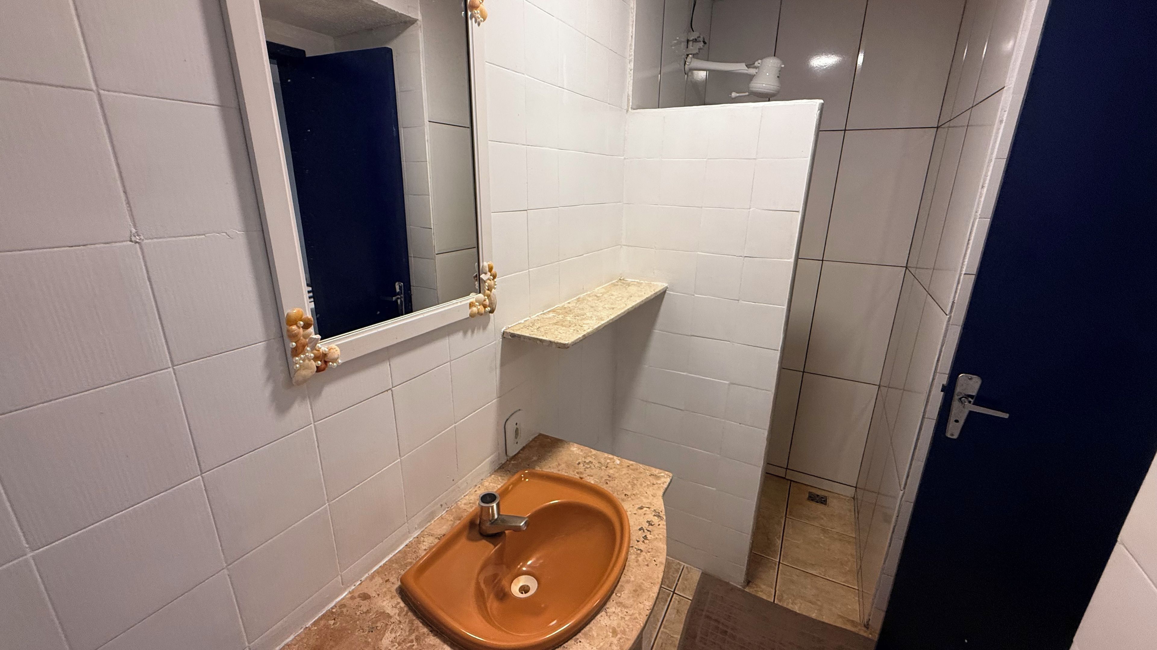 Une salle de bain avec des murs recouverts de carreaux blancs, un lavabo marron, un miroir avec un cadre en bois et une porte bleue.
