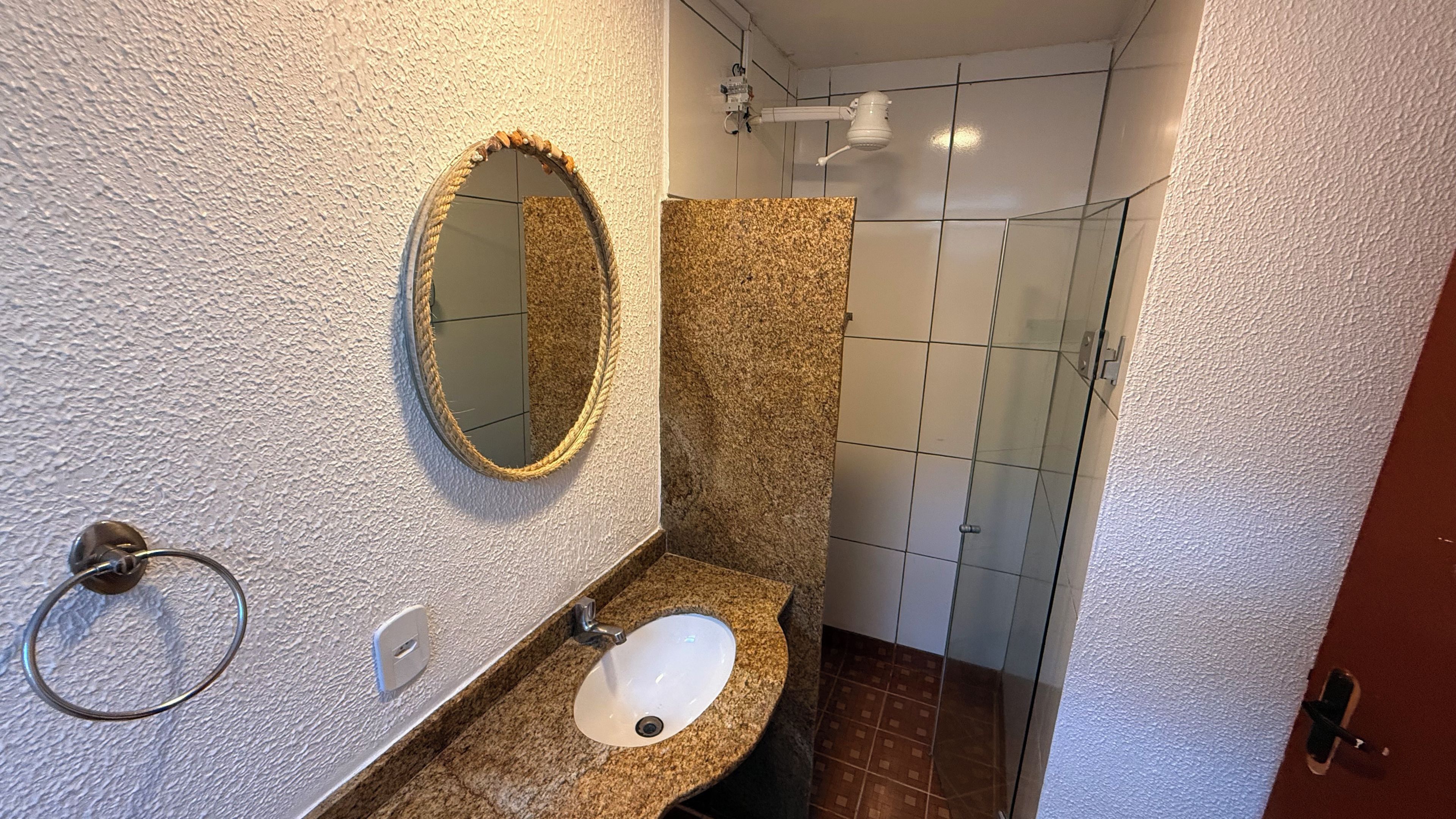 Une salle de bain dotée d’un comptoir en granit, d’un lavabo blanc, d’un miroir rond encadré d’une corde et d’une cabine de douche en verre.