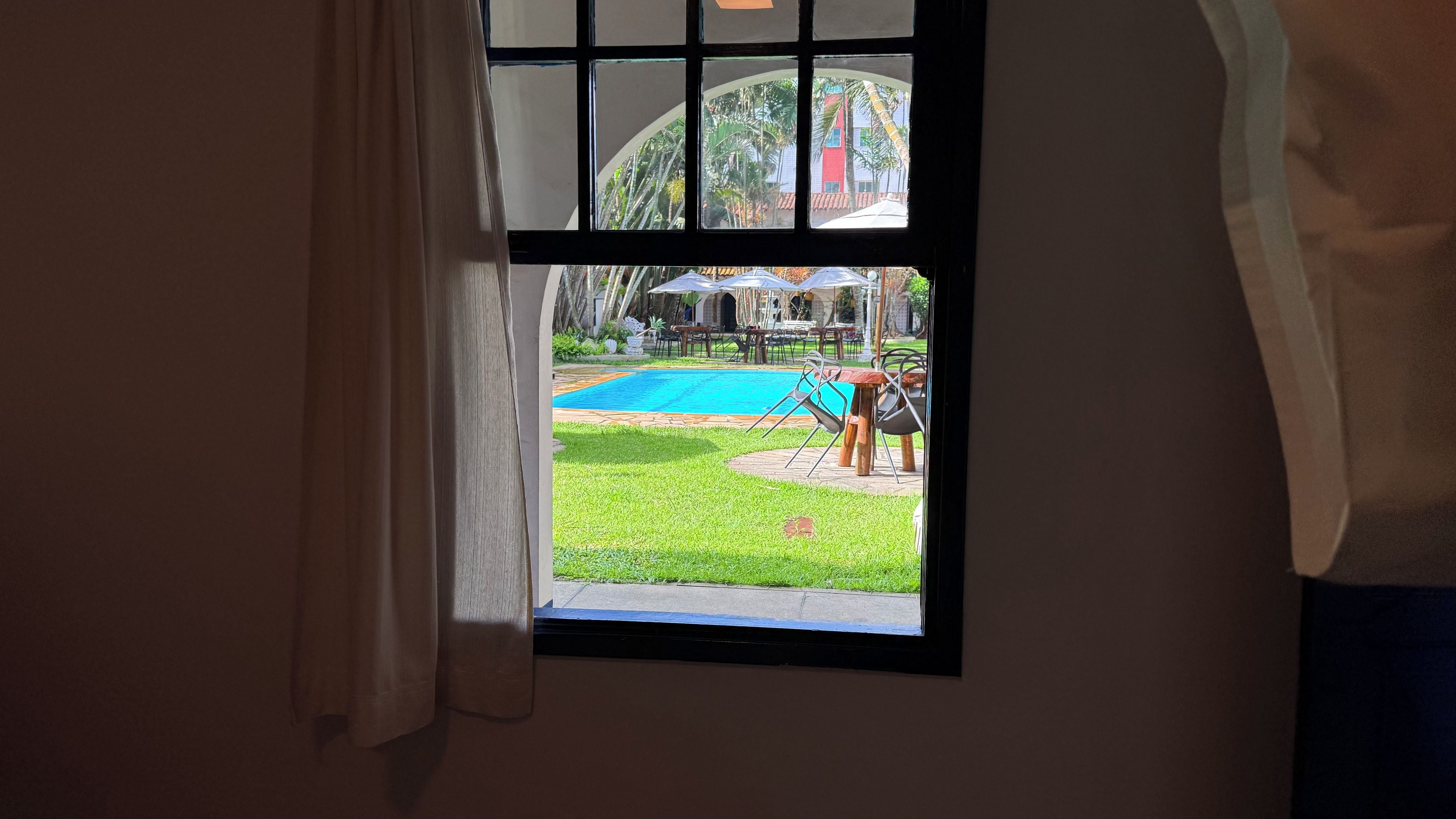 A window with a view of a swimming pool, outdoor seating, and a building with arches.