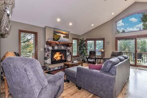 A spacious living room with a stone fireplace, comfortable seating, and large windows offering views of the outdoors. - Eagle River Chain – Steps to Golf & Lake Life! (Eagle River)
