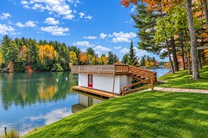 A lakeside cabin with a wooden deck and a red door, surrounded by autumn foliage and a clear sky. - Eagle River Chain – Steps to Golf & Lake Life! (Eagle River)