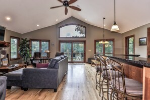 A spacious living area with a large window, a ceiling fan, and a bar area with stools. - Eagle River Chain – Steps to Golf & Lake Life! (Eagle River)