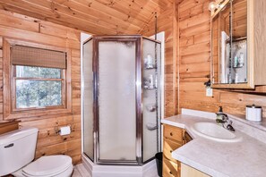 Une salle de bain rustique, avec des murs en bois, comprenant une douche en verre, des toilettes, un lavabo et une fenêtre.