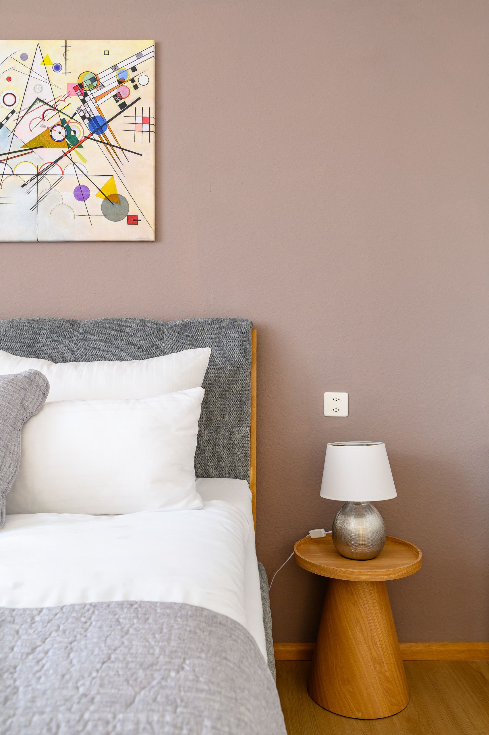Ein Schlafzimmer mit einem Bett, Kissen, einem Nachttisch mit Lampe und einem an der Wand hängenden abstrakten Gemälde.