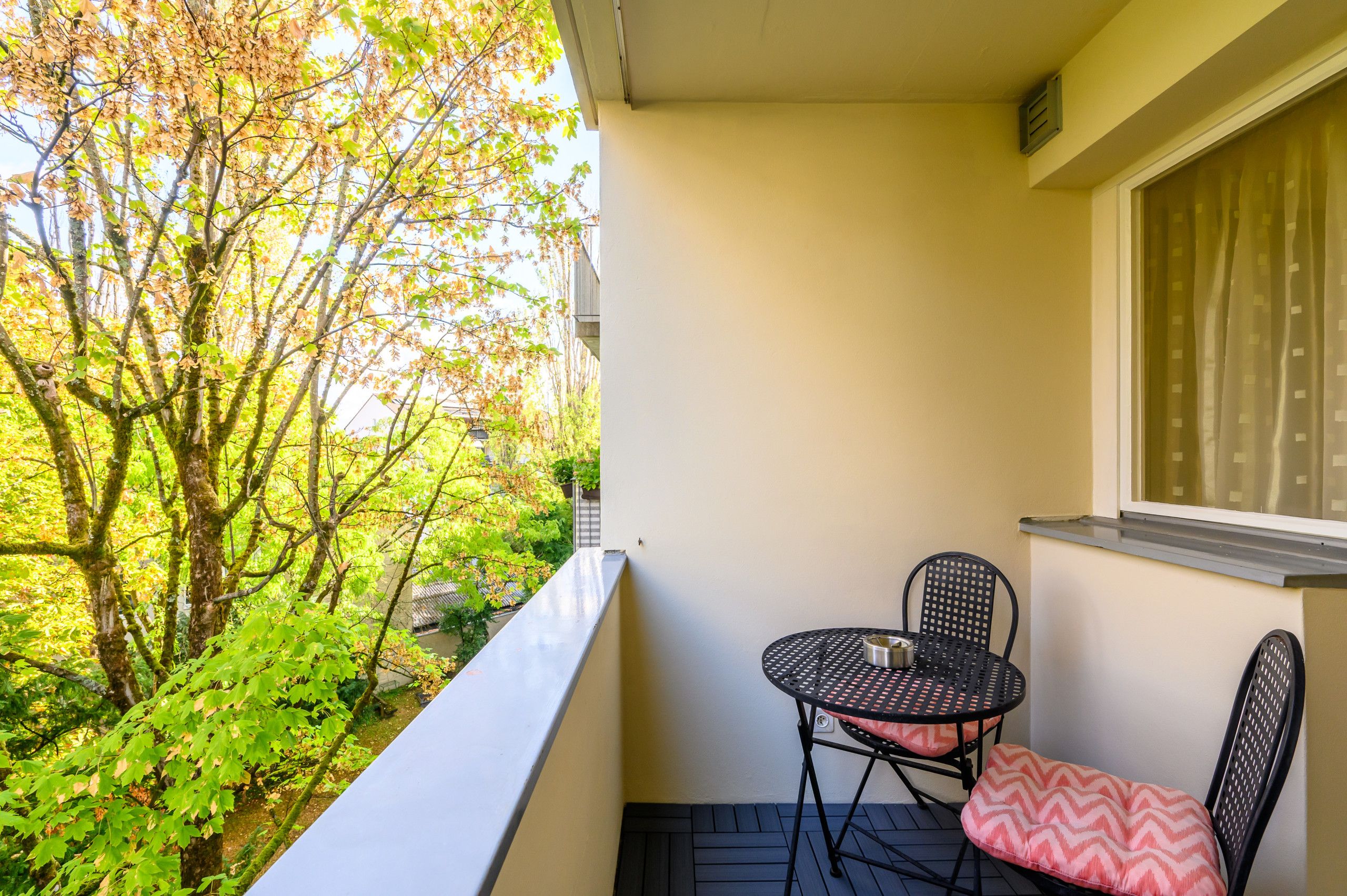 Un balcon avec une table et des chaises, donnant sur un jardin arboré et sur un bâtiment situé au loin.