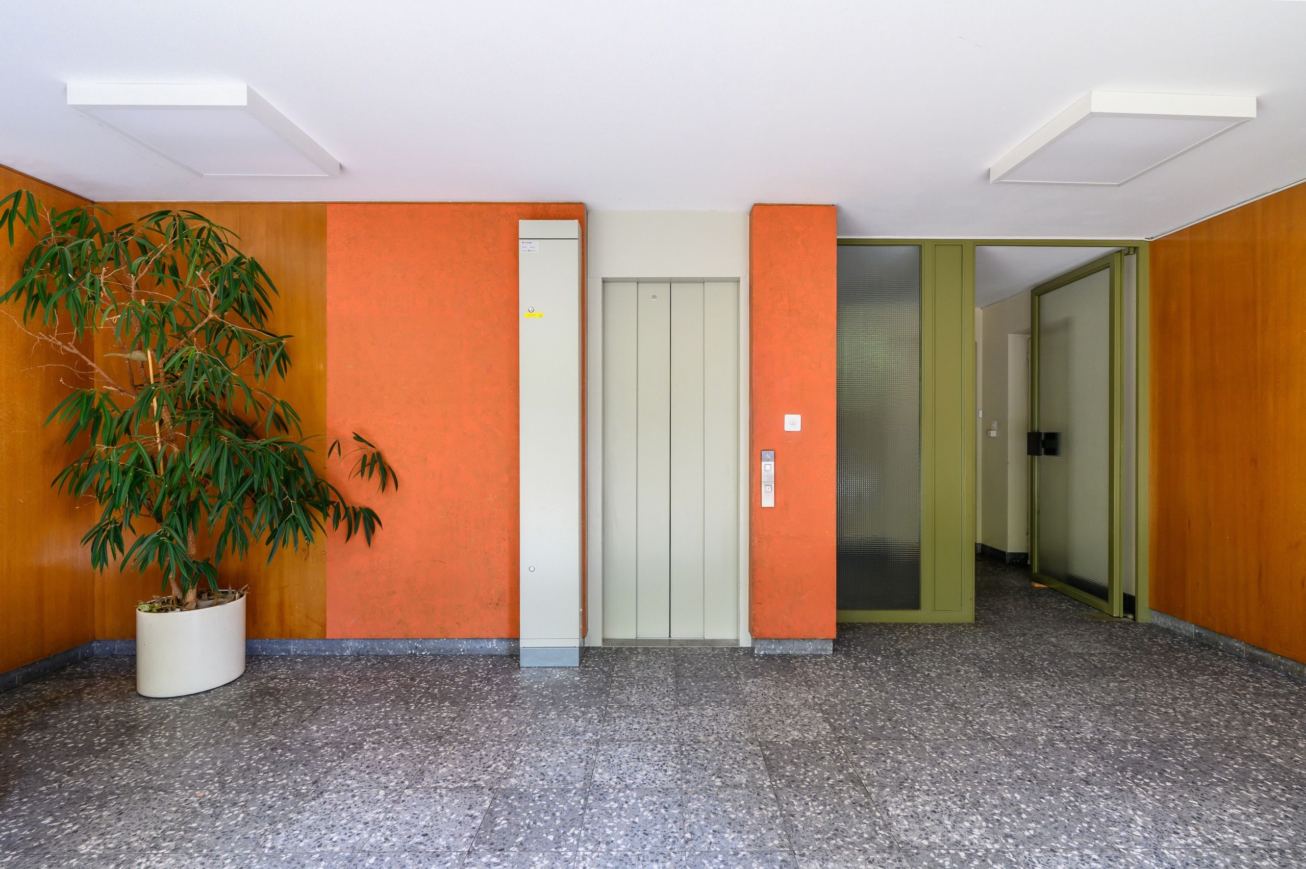 Un couloir avec un pot de plante, une porte avec une pancarte et un mur orné d’un motif texturé.