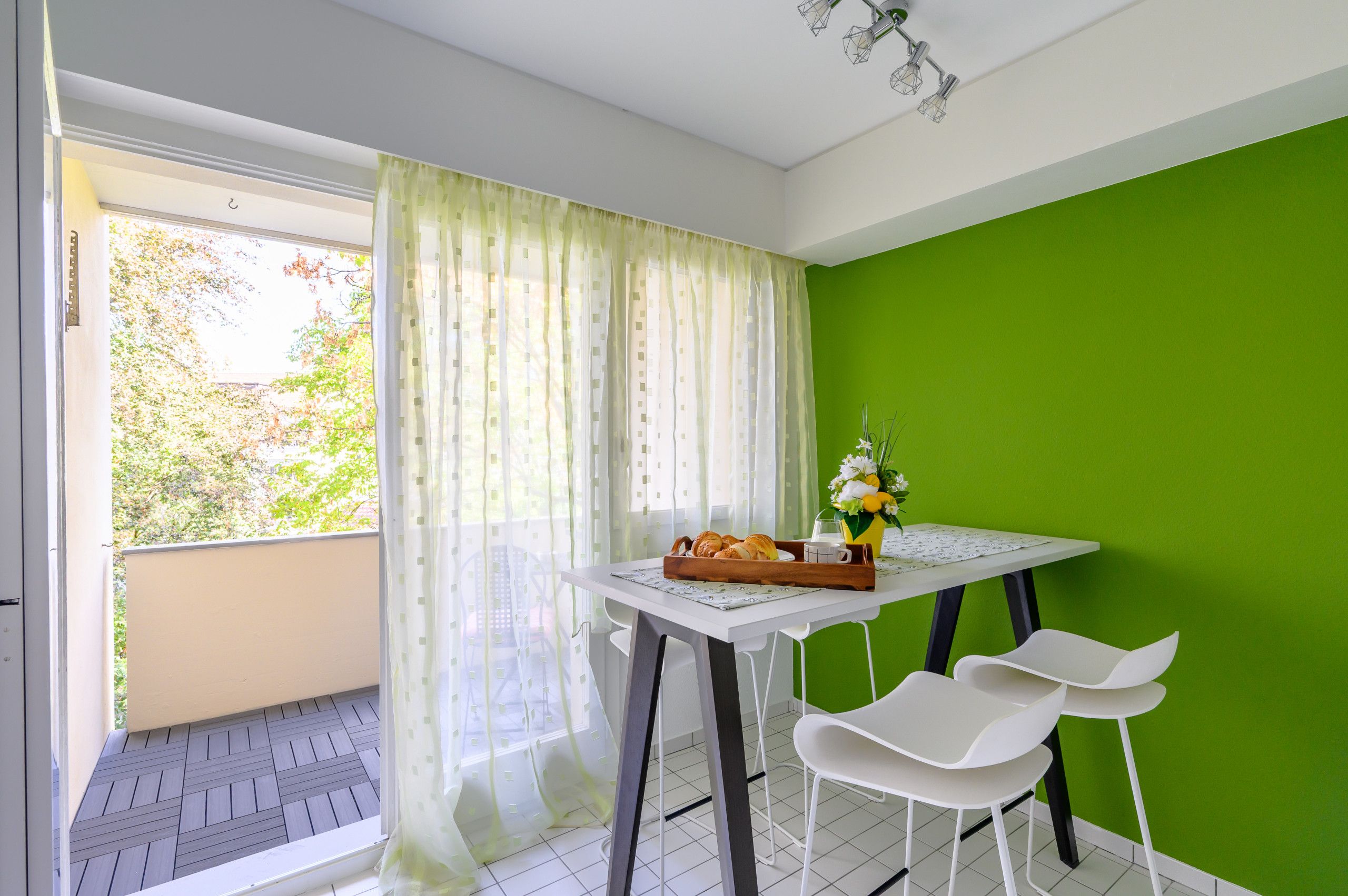 Un balcon avec une table préparée pour le petit-déjeuner, une chaise et une vue sur les arbres à travers la fenêtre.