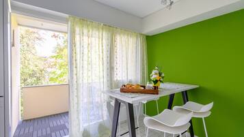 Un balcon avec une table préparée pour le petit-déjeuner, une chaise et une vue sur les arbres à travers la fenêtre.