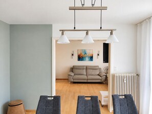 Ein modernes Wohnzimmer mit einer Couch, einem Couchtisch und einem Essbereich mit Stühlen. Ein Heizkörper und ein Gemälde sind an der Wand zu sehen.