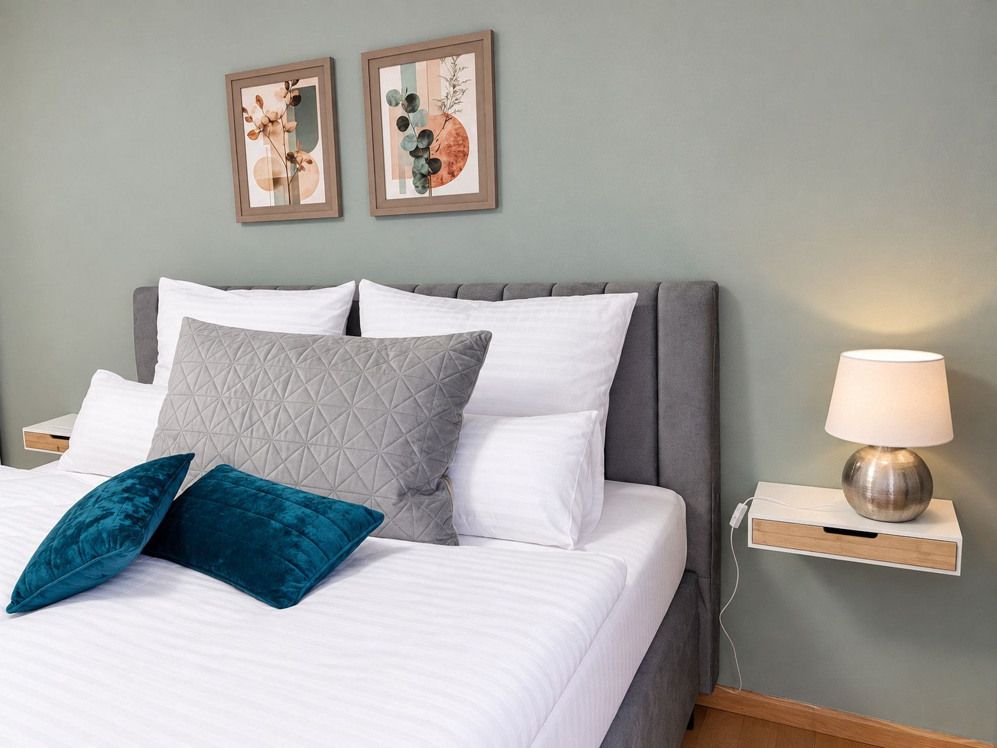Un lit bien fait, avec des oreillers blancs et turquoise, une tête de lit grise et une table de chevet sur laquelle se trouve une lampe.