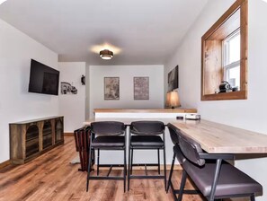 A compact kitchen with a wooden countertop, two black bar stools, a wooden bench, and a TV mounted on the wall. - Sip, Stroll & Stay | Downtown Eagle River Apt (Eagle River)