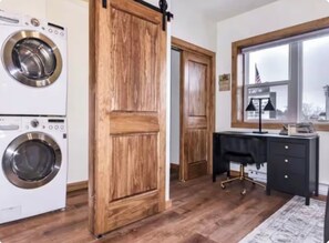 A laundry room with two front-loading washing machines, a wooden sliding door, a desk with drawers, and a window with a view of a flag. - Sip, Stroll & Stay | Downtown Eagle River Apt (Eagle River)