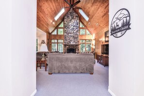 A cozy living room with a stone fireplace, wooden ceiling beams, and a large window overlooking the forest. - Nestled on the banks of East Bay Little Saint Germain Lake! (St Germain)