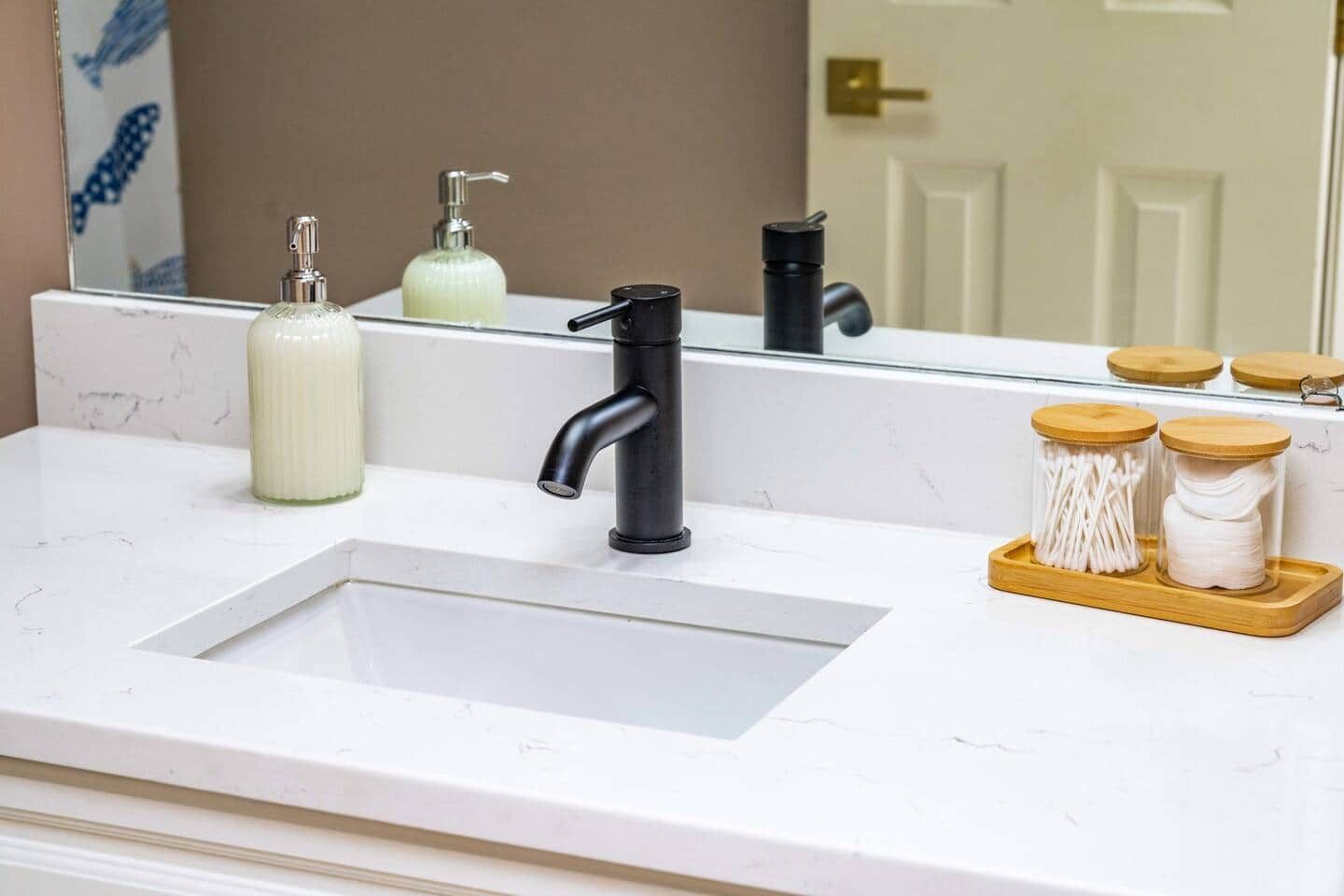 Un lavabo de salle de bain avec un robinet noir, un distributeur de savon et deux pots contenant des cotons-tiges, le tout posé sur un plateau en bois.