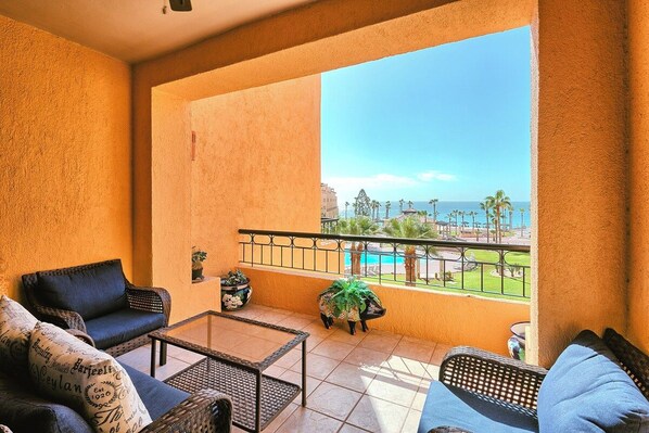Ein Balkon mit Blick auf einen Pool und Palmen, ausgestattet mit einer Couch, einem Couchtisch und einer kleinen dekorativen Schafstatue.