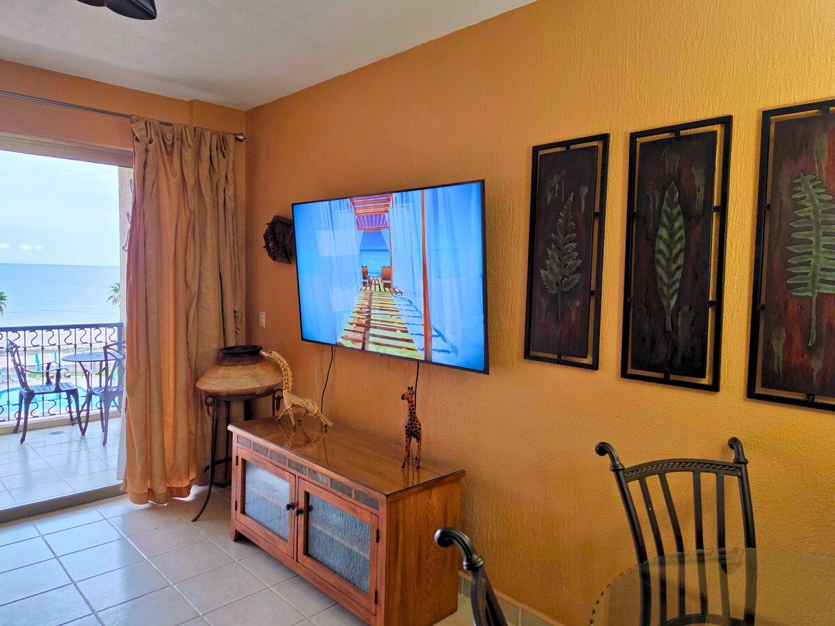 Ein Zimmer mit einem an der Wand befestigten Fernseher, einem Holzschrank, einem Stuhl und einem Balkon mit Stühlen und einem Tisch.