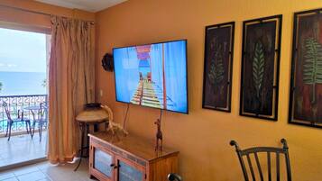 Ein Zimmer mit einem an der Wand befestigten Fernseher, einem Holzschrank, einem Stuhl und einem Balkon mit Stühlen und einem Tisch.