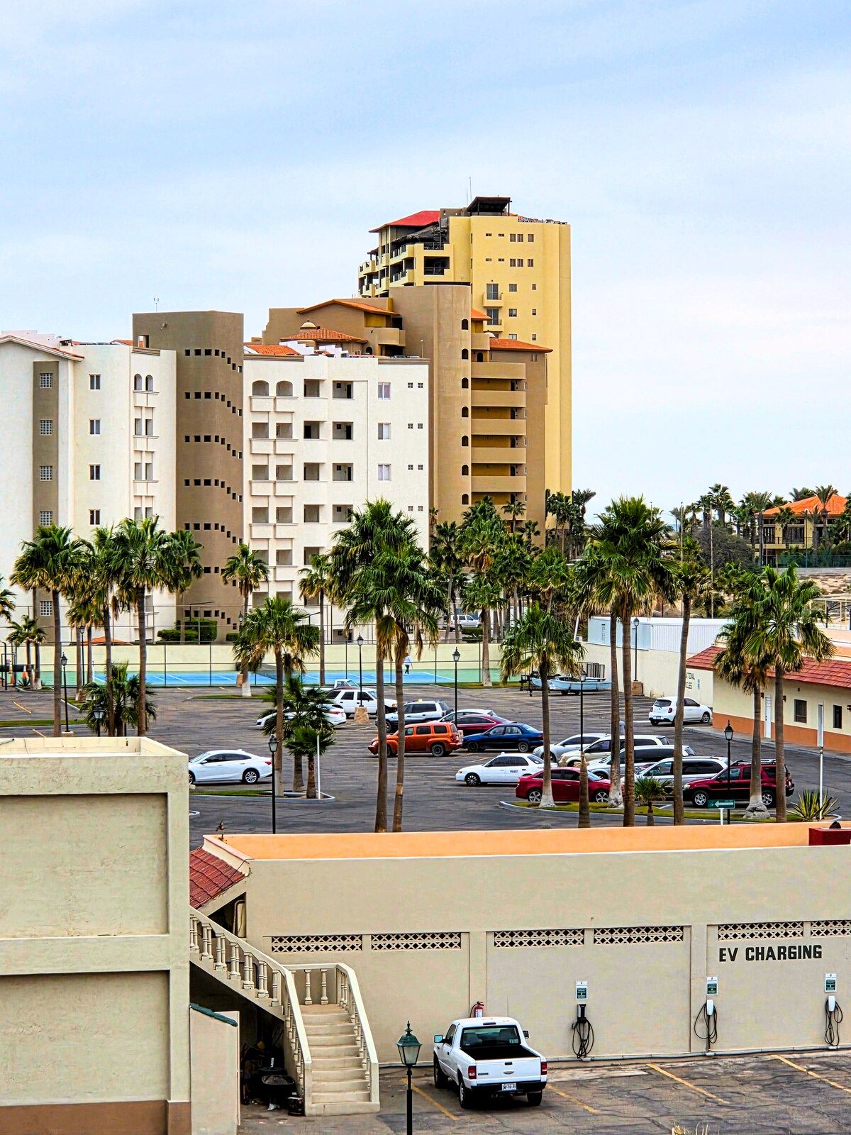 A parking lot with multiple cars, palm trees, and a building with an EV charging station.