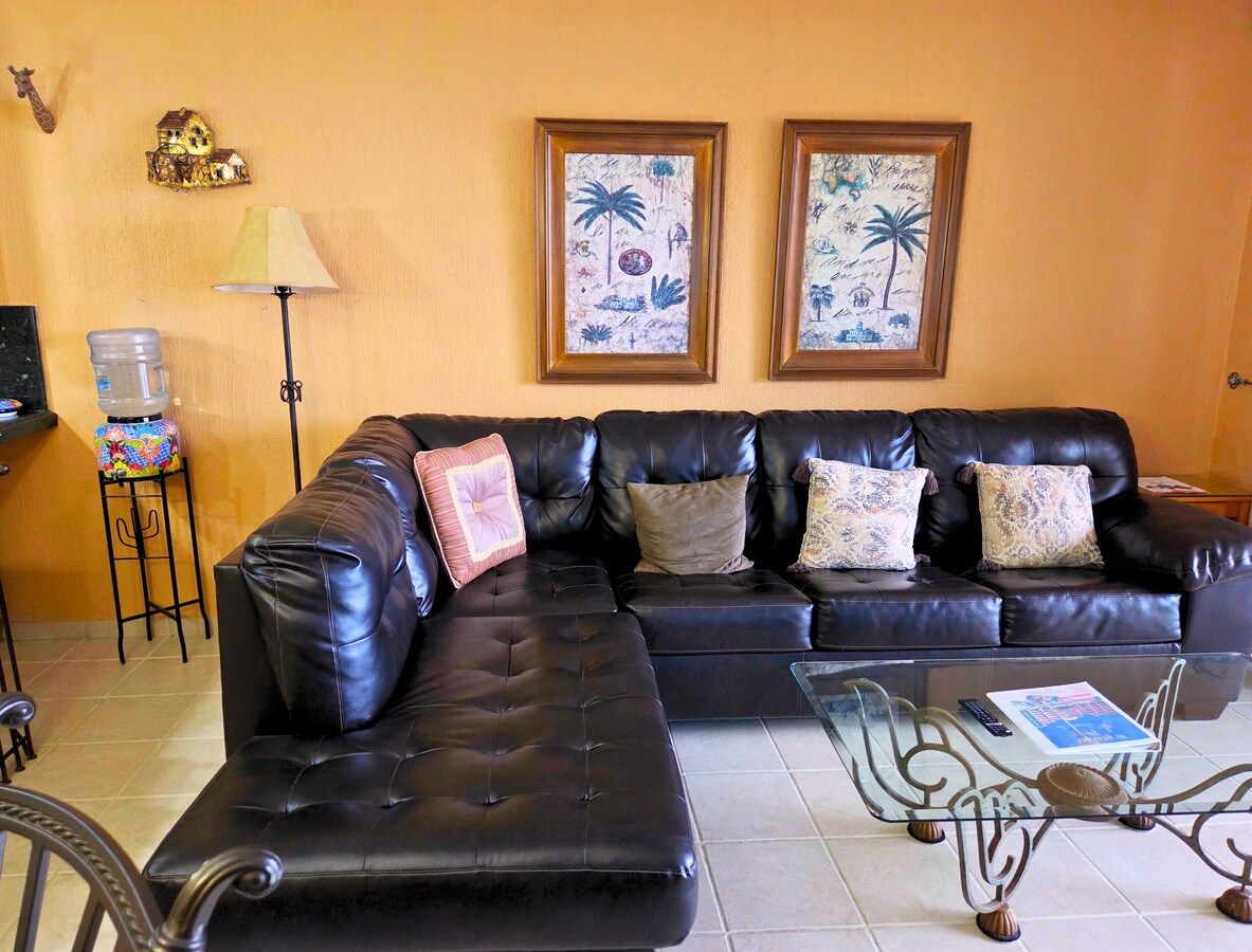 The living room offers a warm and inviting space designed for comfort and relaxation. It features a large black leather sectional sofa accented with decorative pillows, perfect for lounging after a day at the beach.