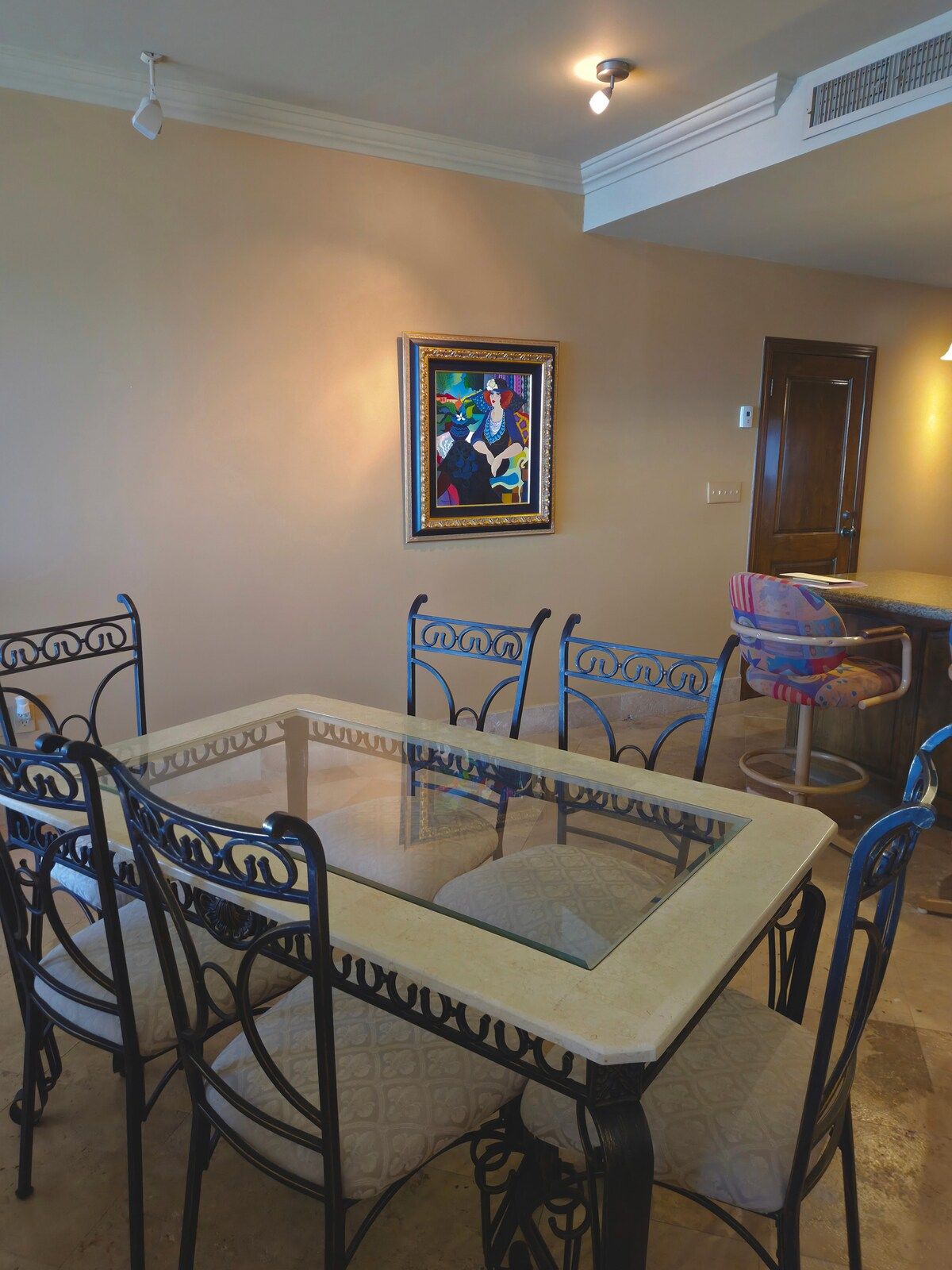 A dining area with a glass-top table, metal chairs, a framed artwork on the wall, and a door in the background.