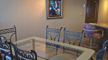 A dining area with a glass-top table, metal chairs, a framed artwork on the wall, and a door in the background.