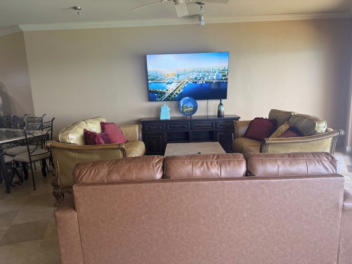 A living room with a brown leather sofa, a television mounted on the wall, and a dining area with a table and chairs.