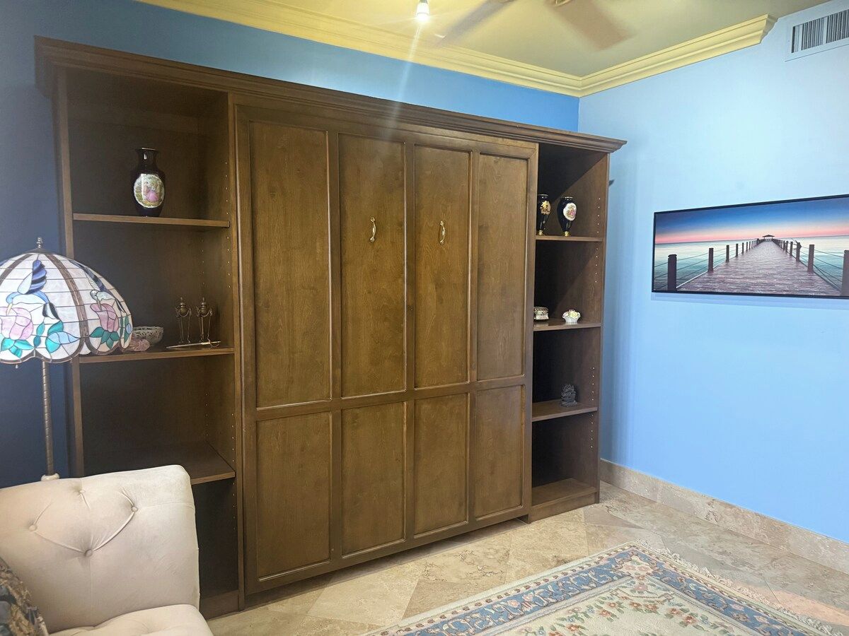 A room with a large wooden wardrobe, a patterned floor rug, and a wall-mounted television displaying a scenic image.