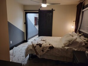 Une chambre à coucher comprenant un lit, un ventilateur de plafond, une fenêtre avec des stores et une porte donnant sur une autre pièce.