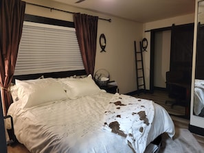 Une chambre à coucher avec un lit, des rideaux, une fenêtre avec des stores, une étagère fixée au mur, une chaise et une échelle.