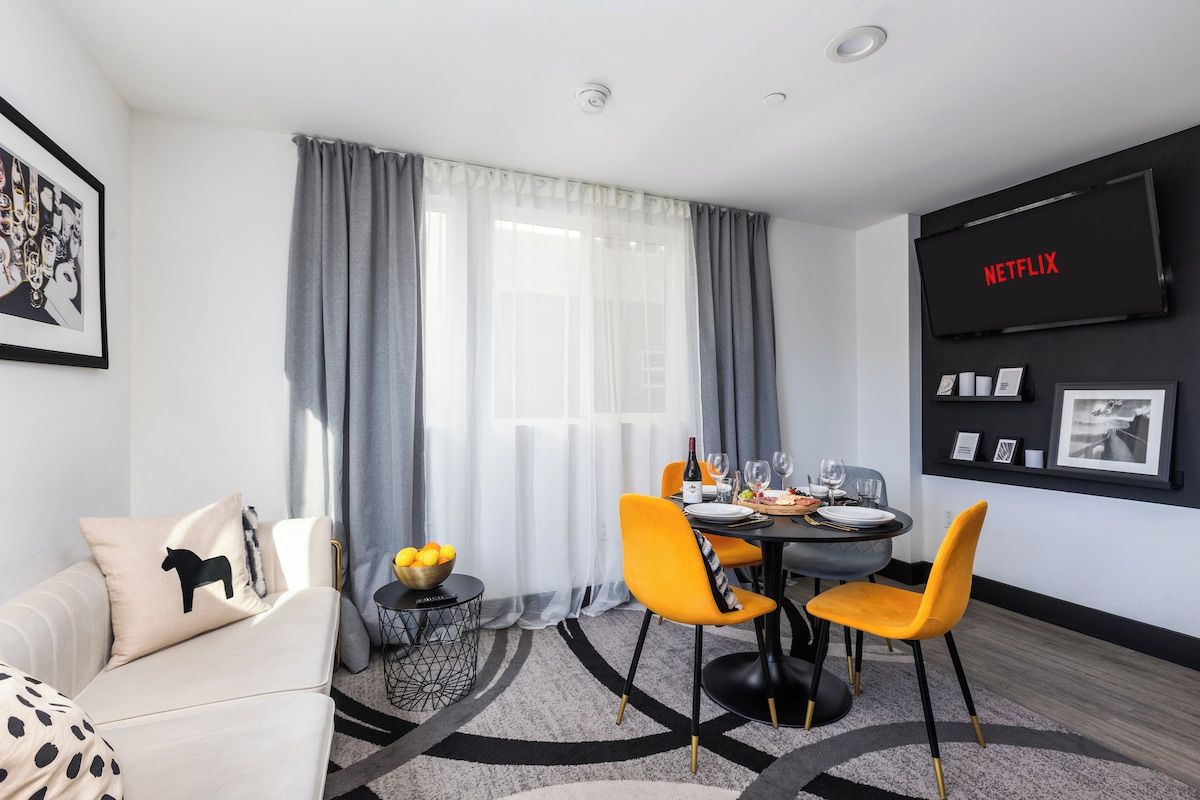 Een moderne woonkamer met een eethoek, een flatscreen televisie en een groot raam met gordijnen.