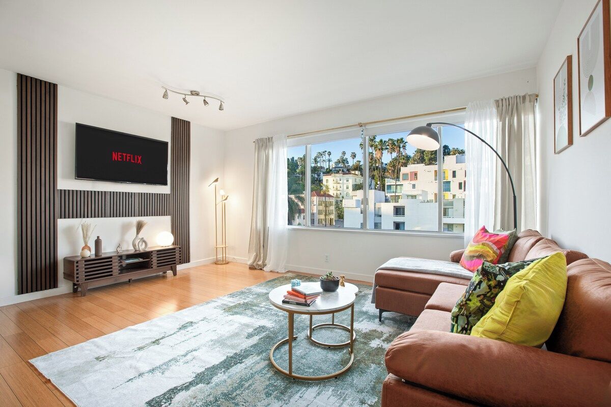 A modern living room with a flat-screen TV, a sofa, a coffee table, and a large window with a view of buildings.