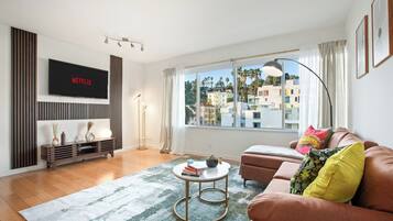 A modern living room with a flat-screen TV, a sofa, a coffee table, and a large window with a view of buildings.