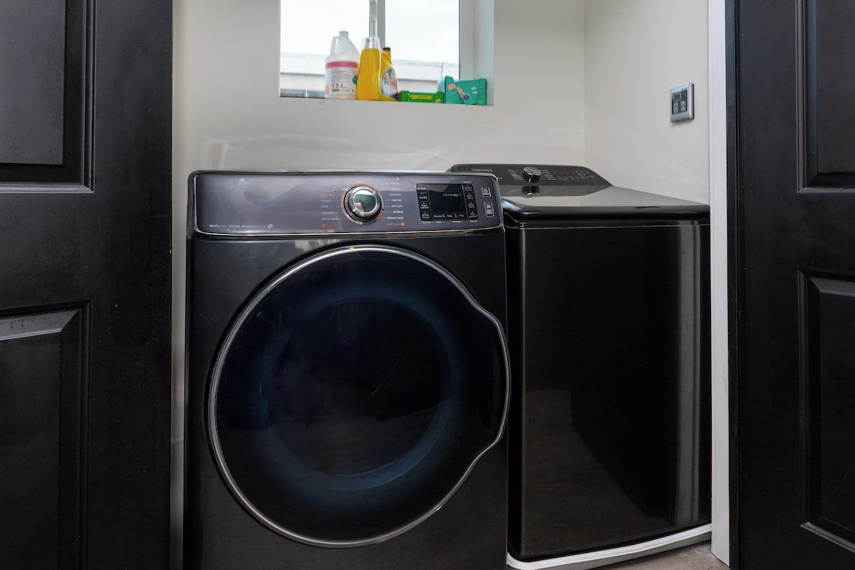 A compact, modern laundry room with a front-loading washing machine and dryer, a small window with a view, and a shelf with cleaning supplies.