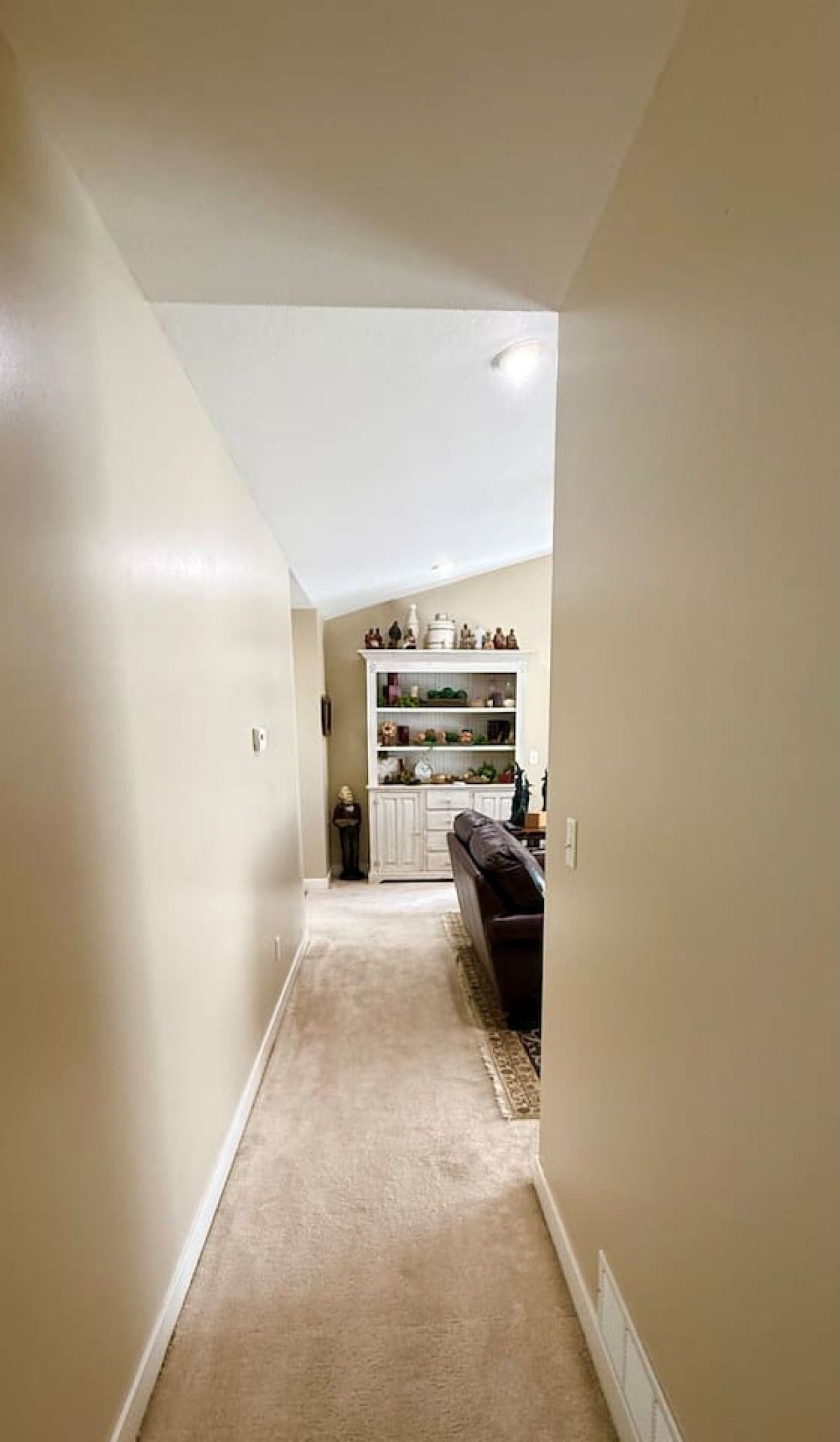 A narrow hallway with a carpeted floor leading to a room with a dark-colored sofa and a white cabinet with decorative items.