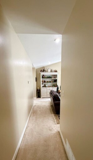 A narrow hallway with a carpeted floor leading to a room with a dark-colored sofa and a white cabinet with decorative items. - Hop Skip & Jump-Good Night Sleep (Boonville)