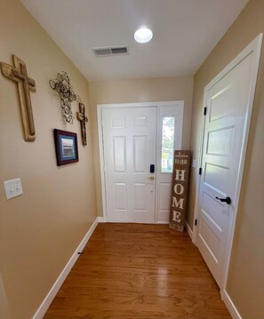 Un couloir doté d’un plancher en bois, de murs blancs et d’une porte blanche avec une fenêtre. On y trouve des éléments de décoration murale, notamment une croix et un tableau encadré.